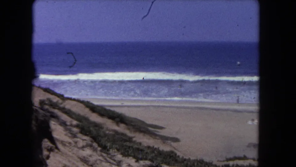 8mm 1962 Los Angeles beach footage shows high surf, a quiet stretch of sand, dune vegetation, and distant swimmers under a blue sky.