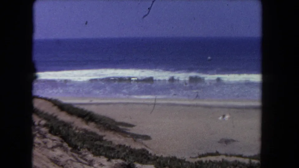Los Angeles beach scene from 1962 with choppy surf, broad sand, low dune plants, and tiny swimmers visible in the ocean.