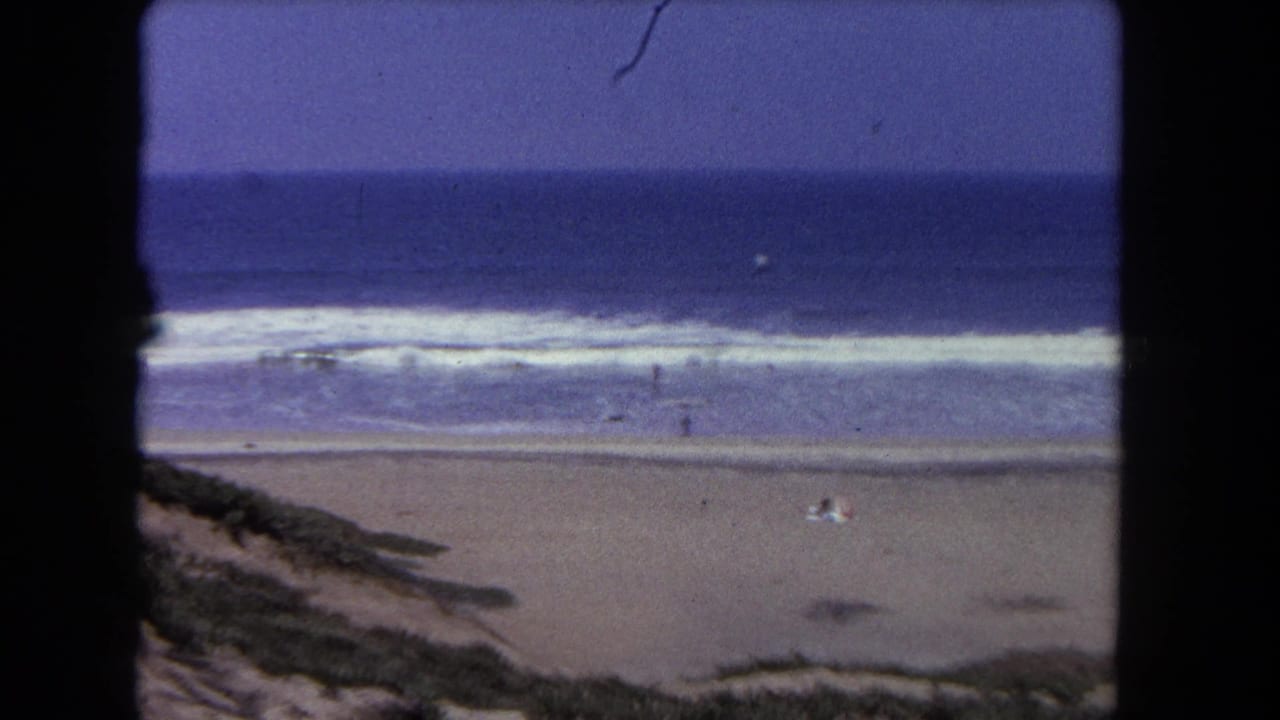 Distant beach with waves, sand, dunes, and a few people seen through dark framing edges.