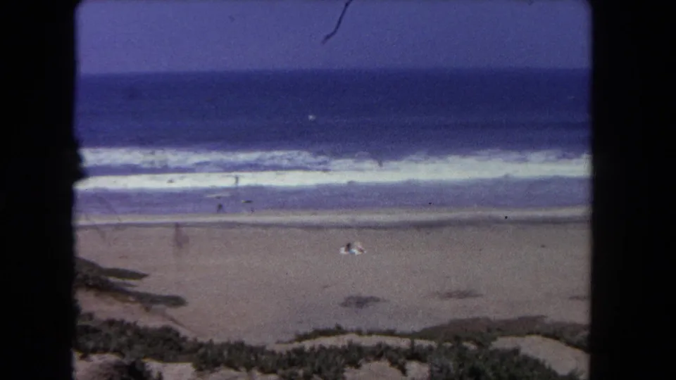 Aged 8mm frame of a Los Angeles beach in 1962, with layered surf, open sand, dune vegetation, and people visible as small distant figures.
