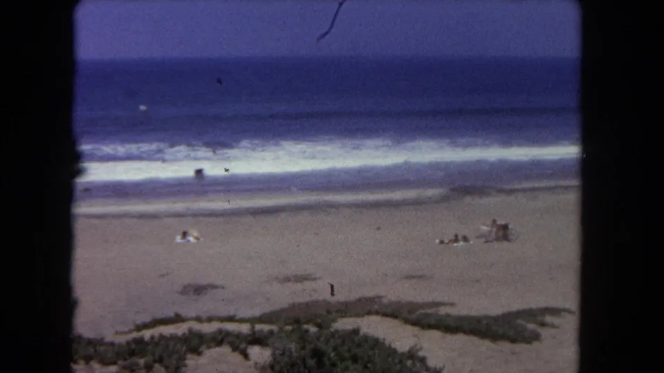 1962 Los Angeles beach footage showing surf, sand, dune vegetation, and several distant people seated or standing near the shoreline.