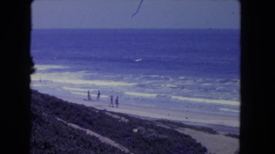 1962 Los Angeles 8mm beach scene with distant walkers along the shoreline, blue ocean surf, sand, and coastal vegetation.
