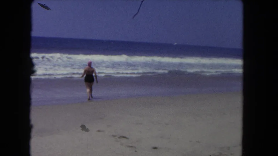 In 1962 Los Angeles beach footage, a woman in a dark swimsuit and pink swim cap runs from the sand toward breaking surf.