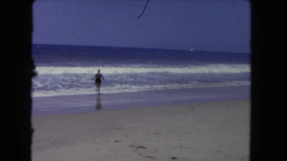 The woman reaches the wet sand near the surf, with the ocean horizon and dark 8mm frame edges visible.