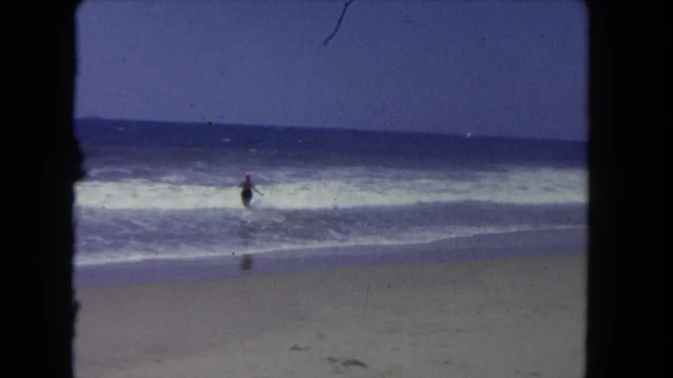 A distant woman wades into the shallow Pacific surf, seen through a soft 8mm home-movie frame.