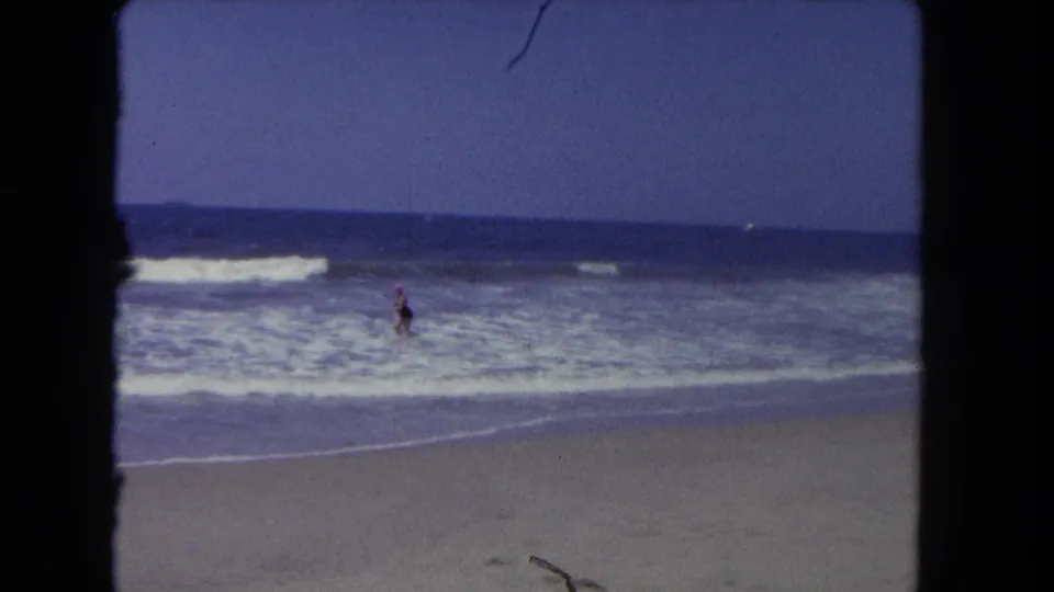 1962 Los Angeles beach footage showing a distant person standing in rolling ocean waves under a pale sky.