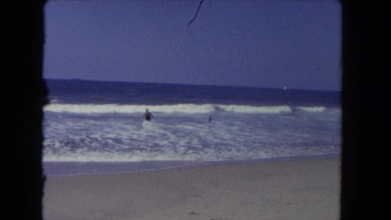 A distant beachgoer wades in Los Angeles ocean surf, framed by black 8mm borders and faded color.