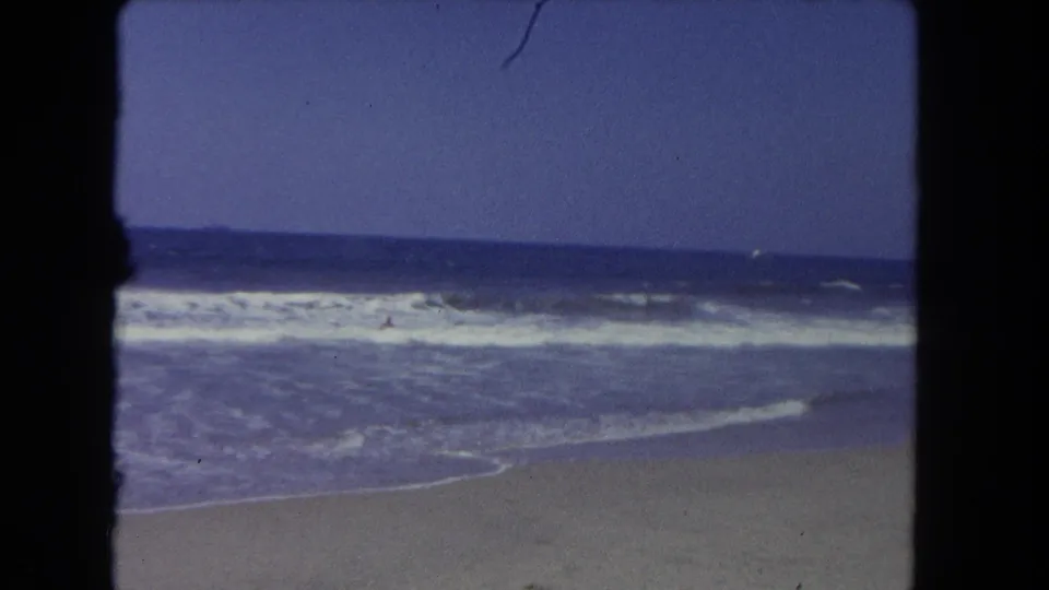 Faded 8mm frame of Los Angeles surf rolling toward shore, with a distant person partly visible in the water.
