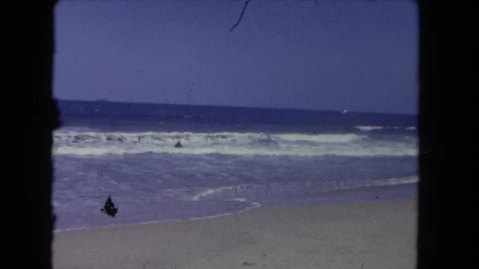 1962 Los Angeles-area beach scene on 8mm film, showing surf, sand, blue water, and a distant person near the shoreline.