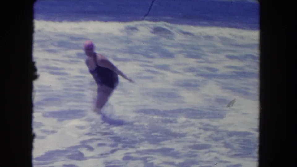 1962 beach home-movie frame showing a woman in a swim cap standing in churning shore water with surf behind her.