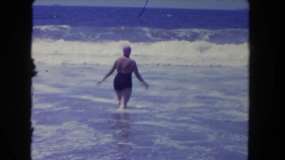 The woman turns away from the camera and walks farther into the surf, framed by blue ocean and foamy breakers.