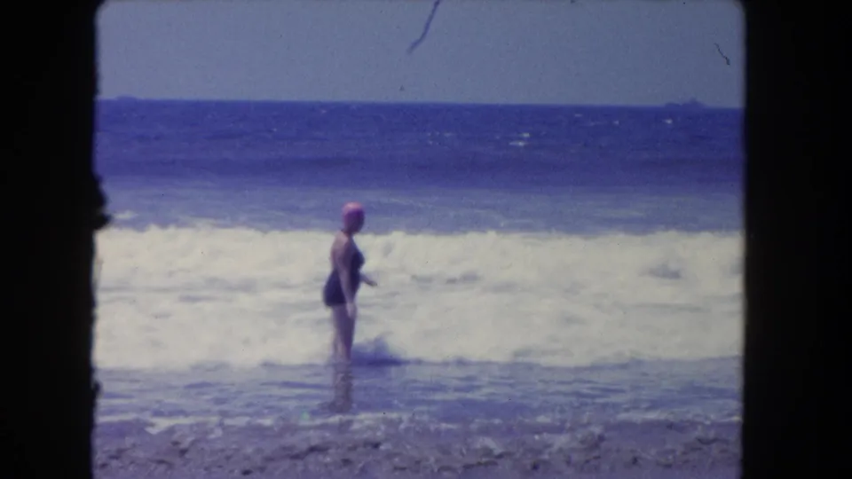 An adult woman in a dark one-piece swimsuit and pink swim cap stands side-on in foamy Pacific surf, with the ocean horizon behind her.