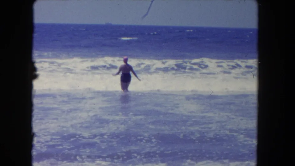 1962 Los Angeles beach footage frame with a woman in a swim cap standing waist-deep near breaking surf.