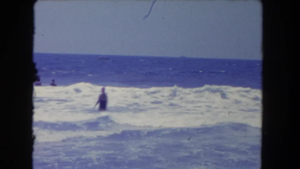 1962 Los Angeles beach home-movie frame showing a woman in the surf with waves, distant swimmers, and a small boat offshore.
