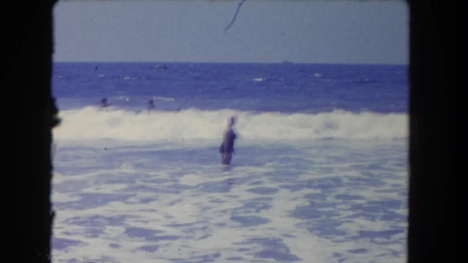 1962 Los Angeles beach frame with a woman in a dark swimsuit standing in rolling surf under a clear sky.