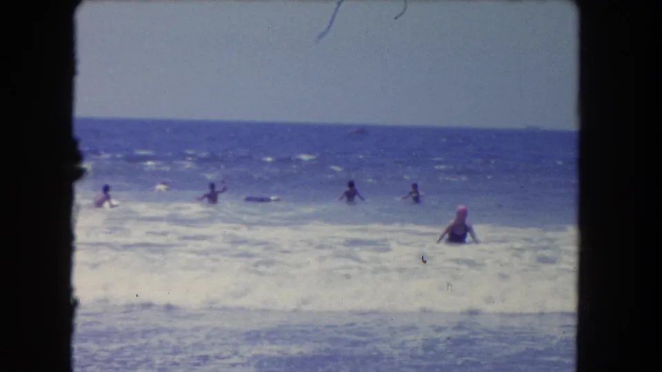 1962 Los Angeles-area beach scene with multiple swimmers in the surf and a woman in a pink cap wading through foreground waves.