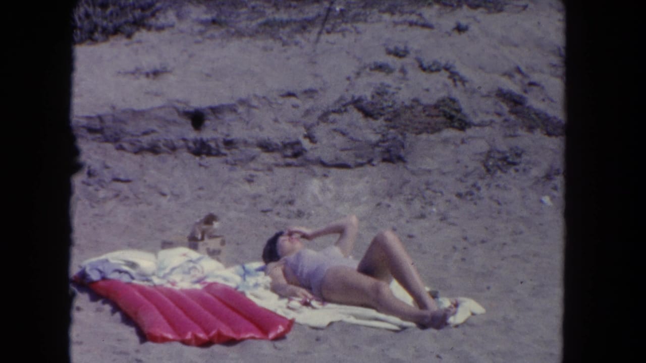A woman sunbathes on a Los Angeles beach in 1962, reclining on towels beside a red mat with dunes behind.