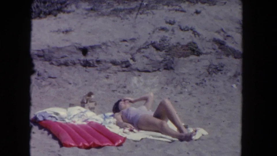 A woman sunbathes on a Los Angeles beach in 1962, reclining on towels beside a red mat with dunes behind.