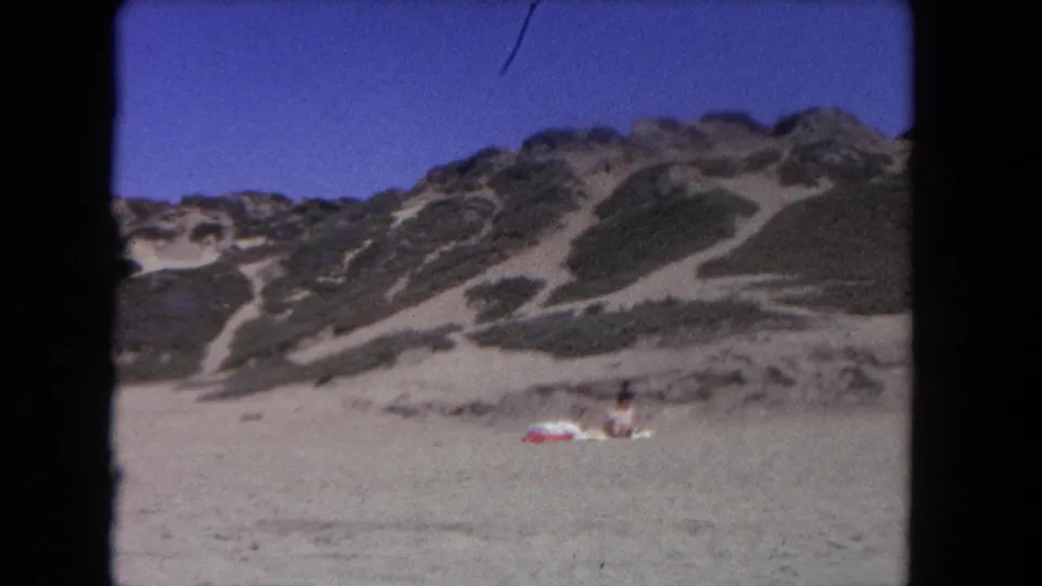 1962 Los Angeles-area beach frame showing dunes, open sand, blue sky, and a distant person seated on the beach.