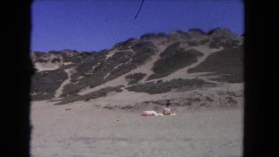 Wide 1962 Los Angeles beach view with low dunes, pale sand, and one distant seated beachgoer under a bright sky.