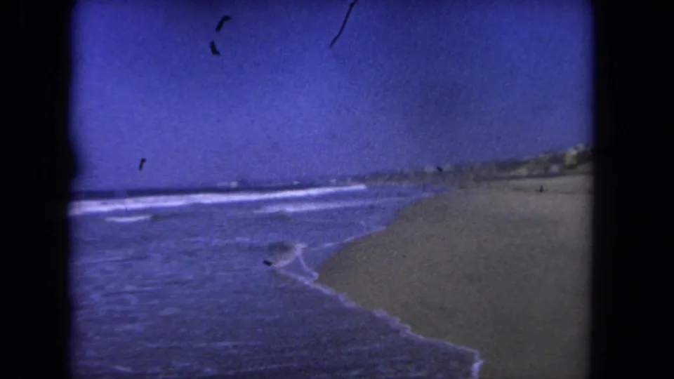 1962 Los Angeles beach scene with surf foam, open sand, distant coastal structures, and visible film debris.