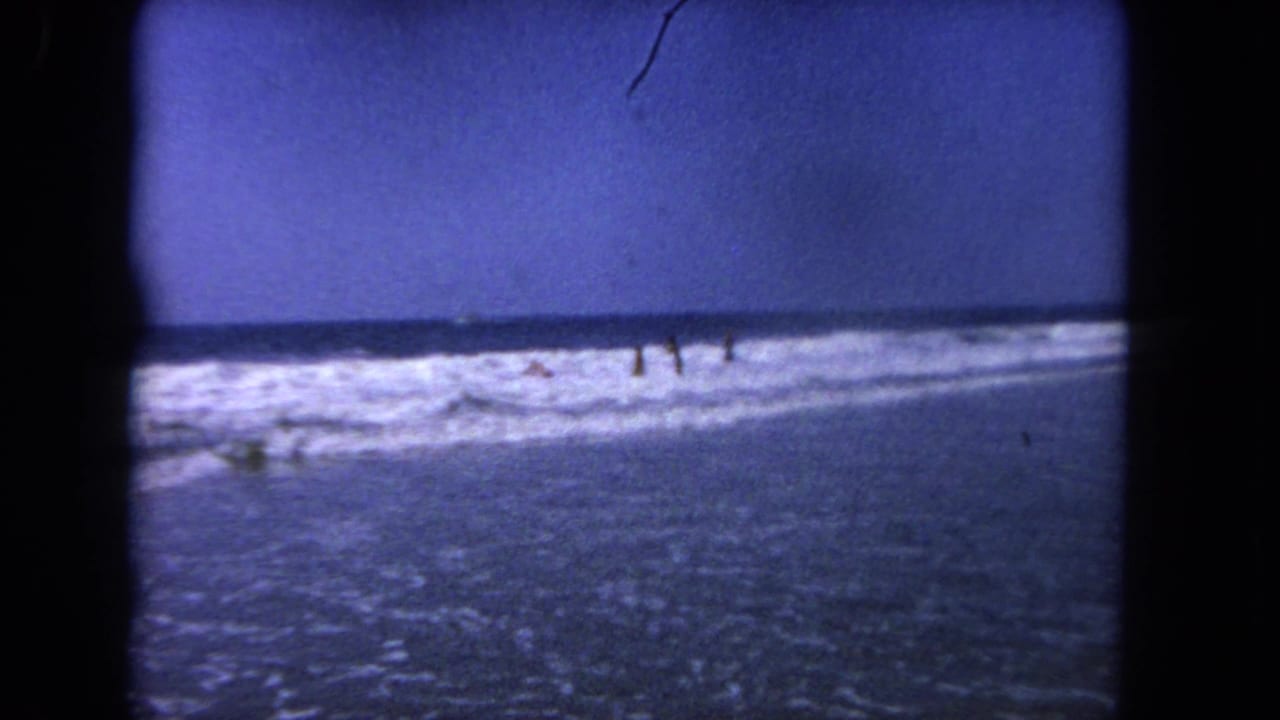 Ocean surf with several distant people standing in shallow waves beneath a blue sky.