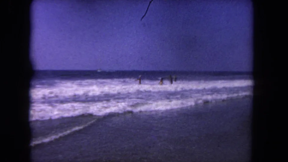 Distant figures remain in the surf as waves break along a 1962 Los Angeles beach scene.