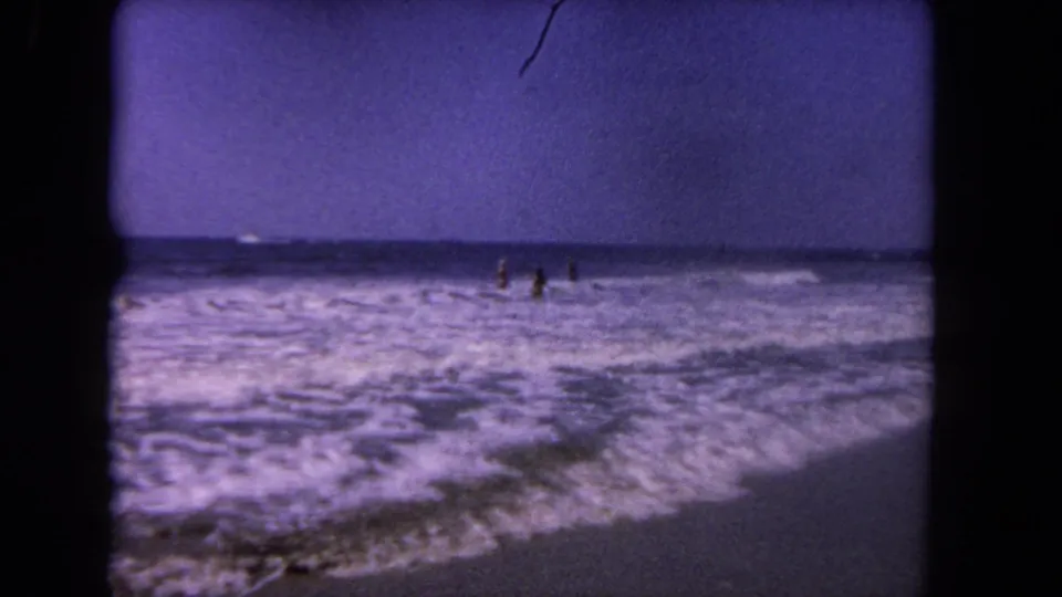 1962 Los Angeles beach footage shows distant figures in shallow surf under a blue-purple sky.