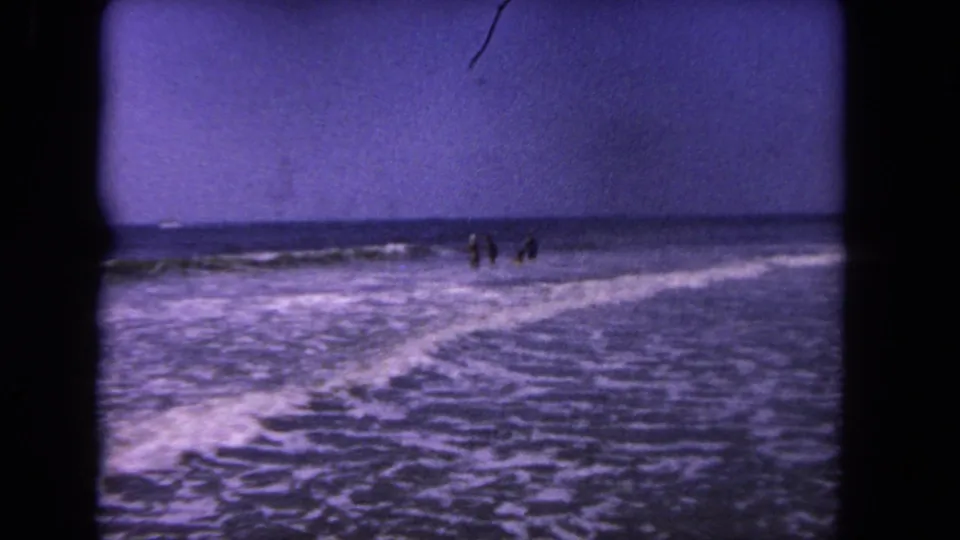 1962 8mm Los Angeles shoreline frame with four distant figures standing amid diagonal lines of surf foam.