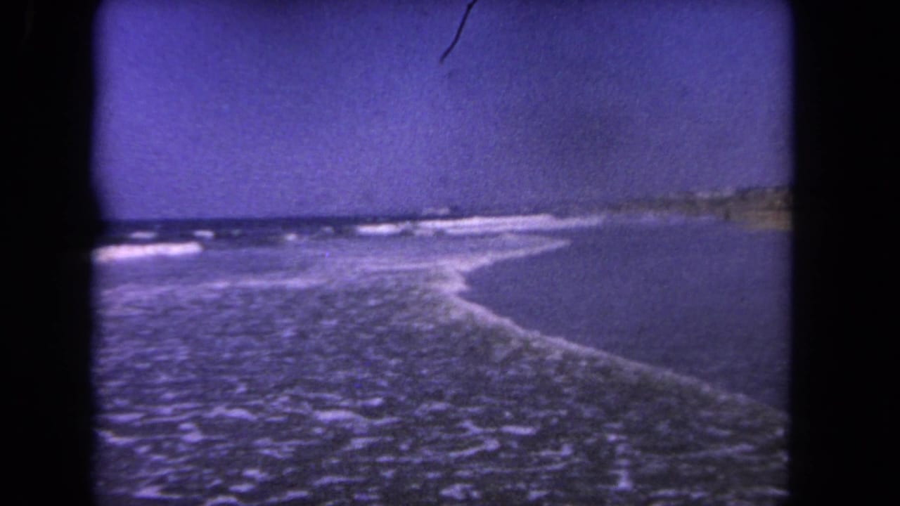 1962 Los Angeles-area beach view with surf breaking diagonally toward shore and faint distant land or shoreline at the horizon.