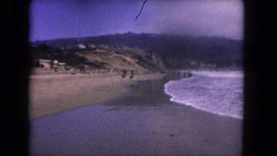 1962 Los Angeles-area beach scene on 8mm film with surf washing onto sand below a low hillside with scattered structures and distant beachgoers.