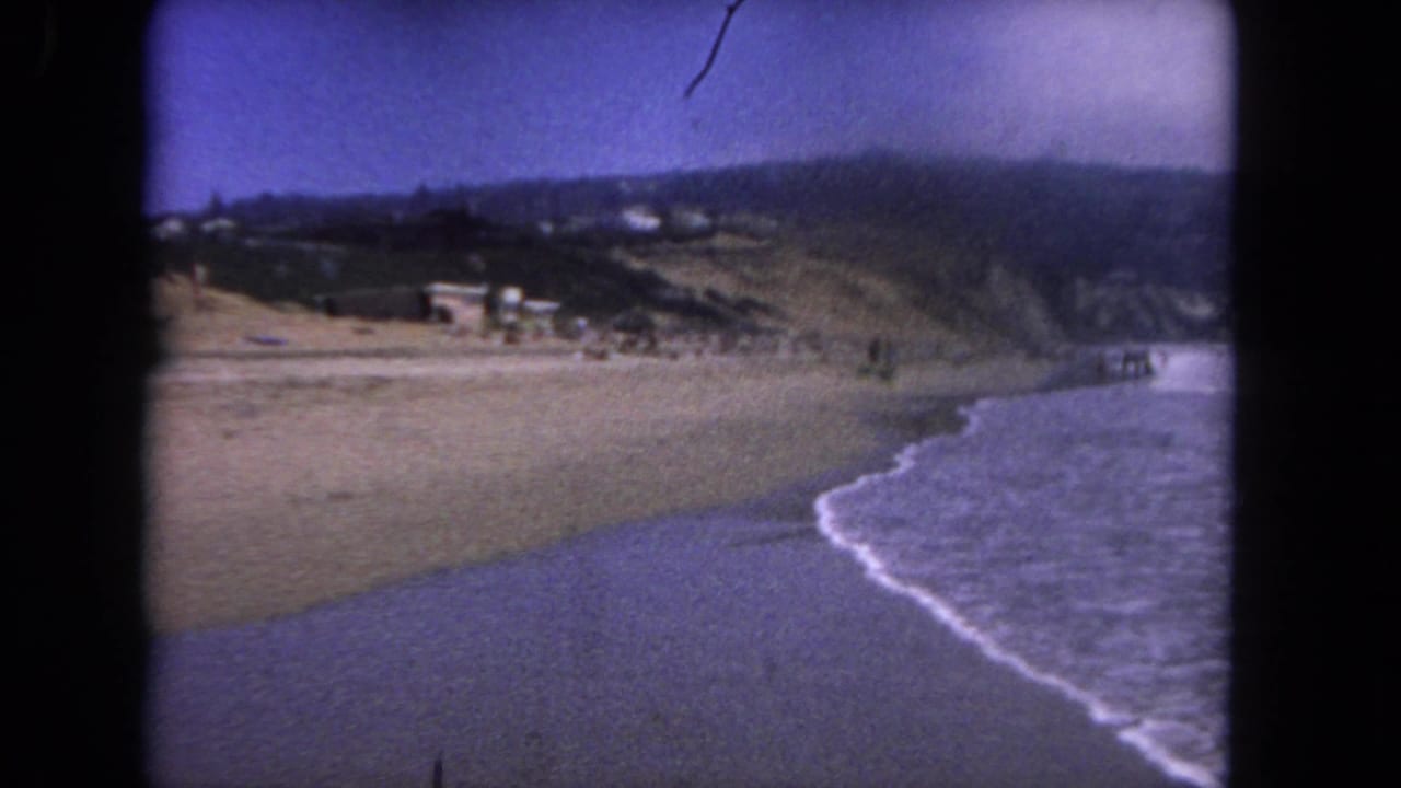 Curving shoreline with ocean surf, open sand, hillside, and small distant figures.