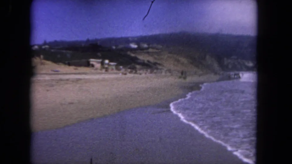 1962 Los Angeles-area coastal view showing a curving sandy shoreline, blue-purple surf, hillside terrain, scattered buildings, and distant people near the waterline.