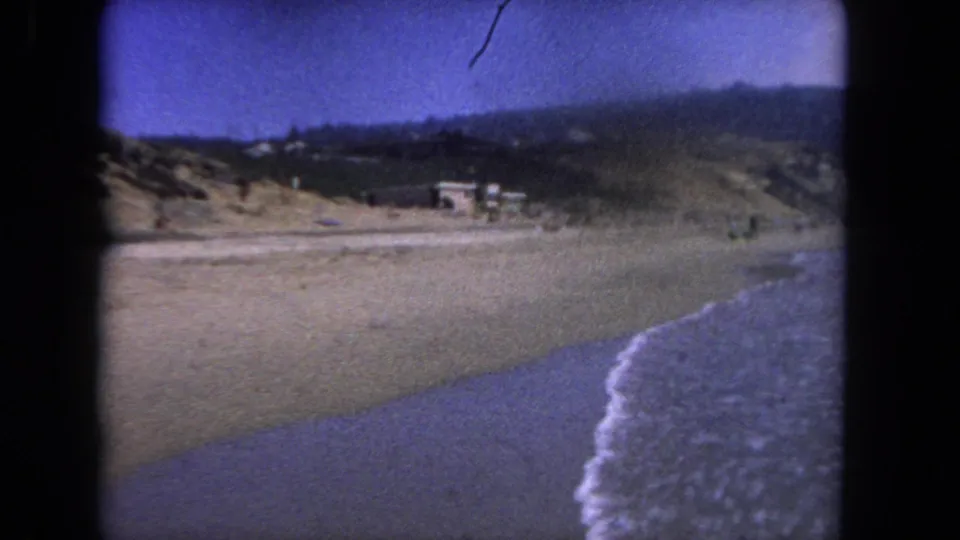 Handheld 8mm frame from a 1962 Los Angeles-area beach, with surf at the right edge, open sand in the foreground, hillside beyond, and small beachgoers in the distance.