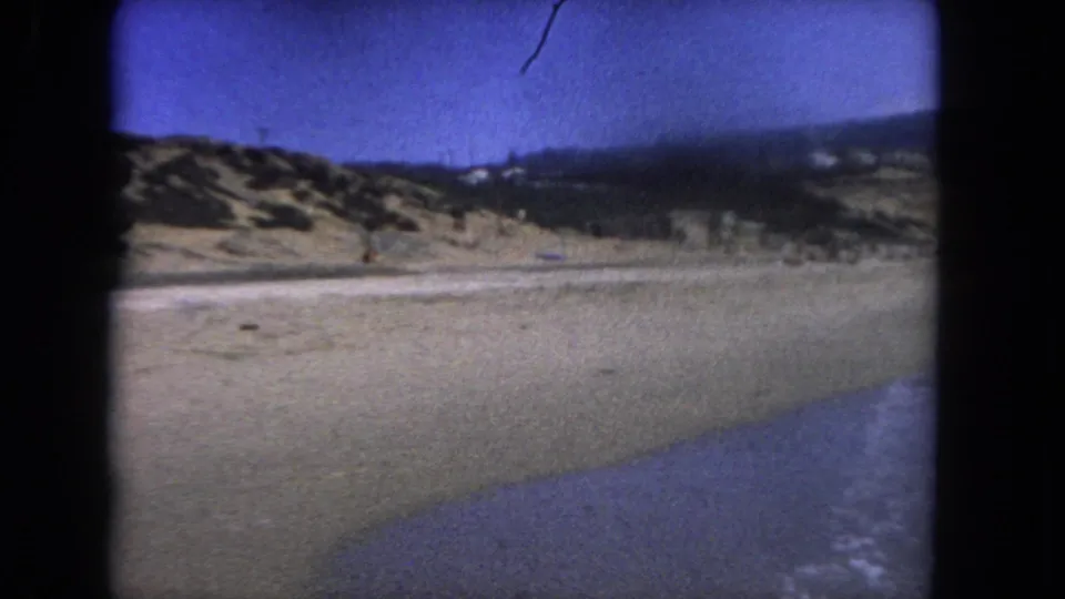 Final 1962 Los Angeles-area beach frame showing pale sand, shallow surf, a low coastal hillside, scattered structures, and distant people within a visible 8mm film gate.