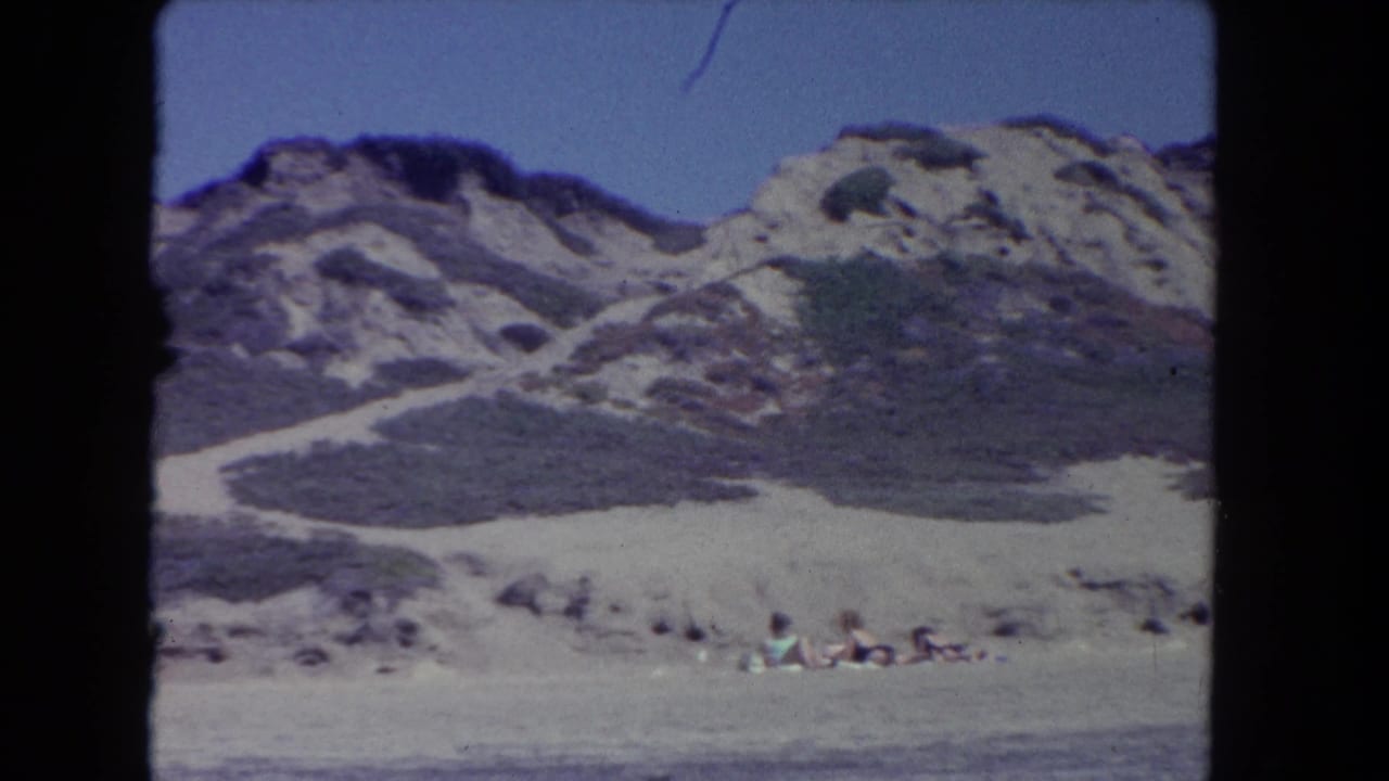 Coastal dunes and rocky hills above a sandy beach with a few distant people.