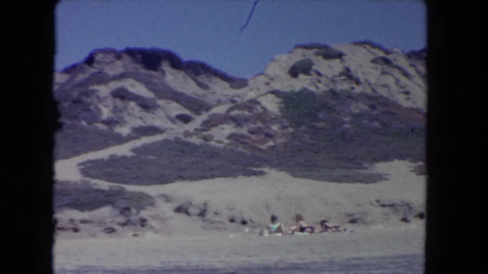 1962 Los Angeles-area 8mm frame showing sandy beach foreground, scrub-covered dunes, rocky ridges, blue sky, and small distant beachgoers.