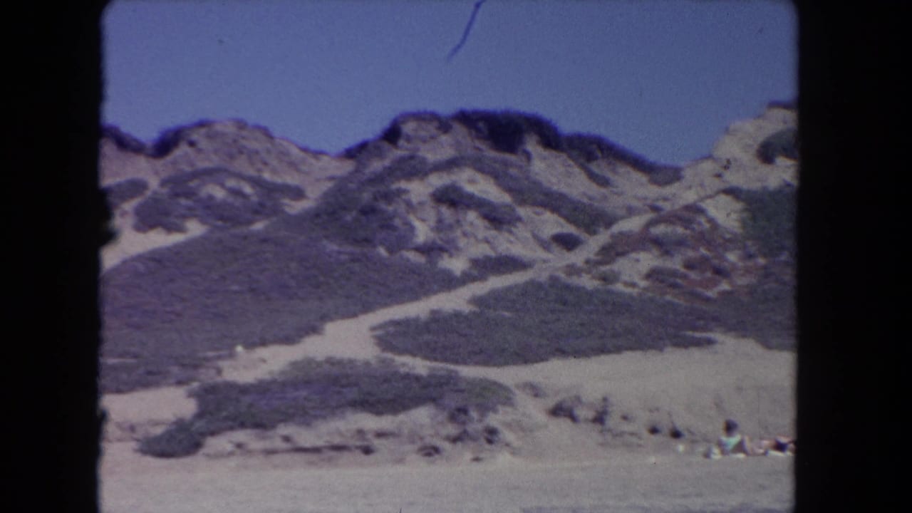Scrub-covered sandy hillside with rocky ridges and distant beachgoers at the lower right.