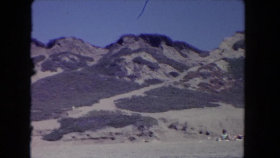 1962 Los Angeles-area coastal landscape with pale sand paths, low shrubs, rocky hilltops, blue sky, and small beachgoers near the edge.