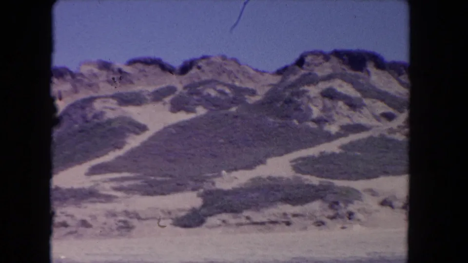Soft 8mm frame from 1962 Los Angeles showing scrub-patterned dunes, rocky upper slopes, pale beach foreground, and no clearly identifiable landmarks.