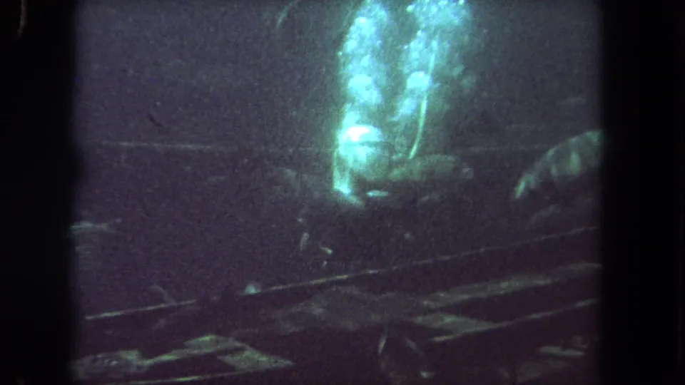 1962 Los Angeles 8mm frame showing a helmeted diver in dark aquarium water with bubbles above and fish silhouettes around the tank.