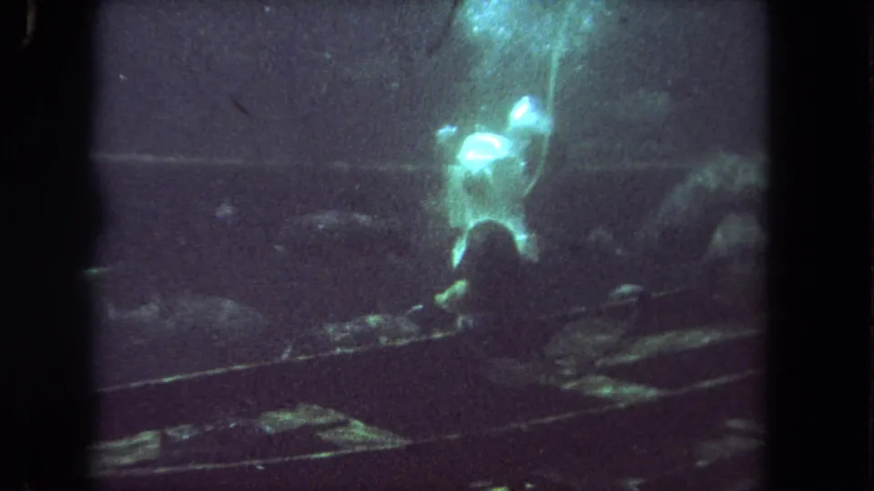 1962 Los Angeles-area 8mm frame showing a scuba diver near sunken boat wreckage with fish in green murky water.