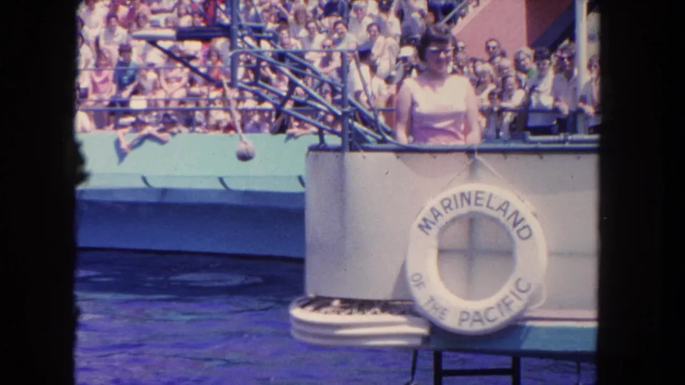 1962 home-movie frame at Marineland showing the platform area, woman at the rail, branded life ring, pool water, and audience.