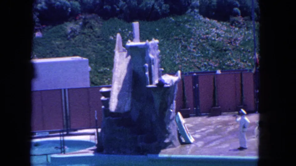 Wide 1962 Marineland frame showing the poolside rock structure, red wall and a white-uniformed trainer on the stage.