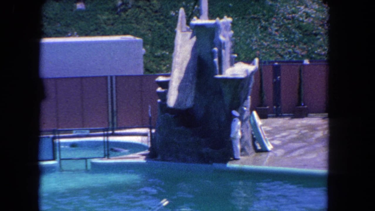 1962 Los Angeles home-movie frame with a homemade robot figure, pool, slide structure, and a distant person in white.