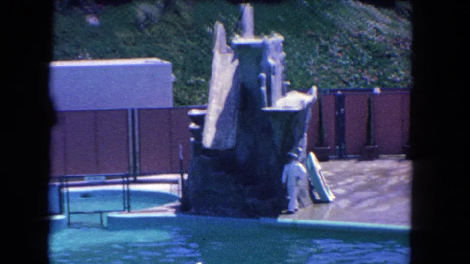 1962 Los Angeles home-movie frame with a person moving beside a homemade robot figure, pool water, deck, and fencing.
