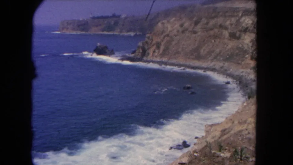1962 home-movie coastal frame showing foamy surf, steep sandy cliffs, and scattered rocks along the Los Angeles shoreline.