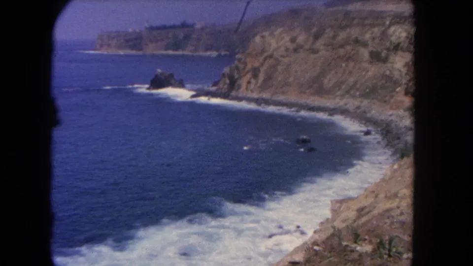 Archival 8mm coastal view from Los Angeles in 1962, with waves moving toward a rugged cliff shoreline and dark frame edges visible.