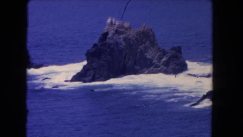 1962 Los Angeles 8mm frame centered on a jagged offshore rock formation with waves foaming around it.