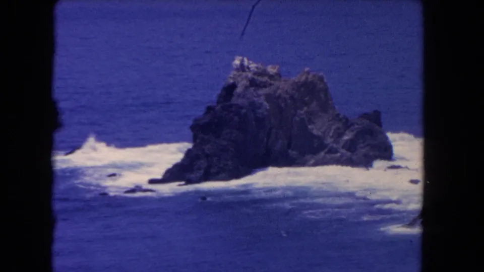 1962 Los Angeles-area coastal home-movie frame showing a rocky outcrop in blue sea with waves breaking around it.